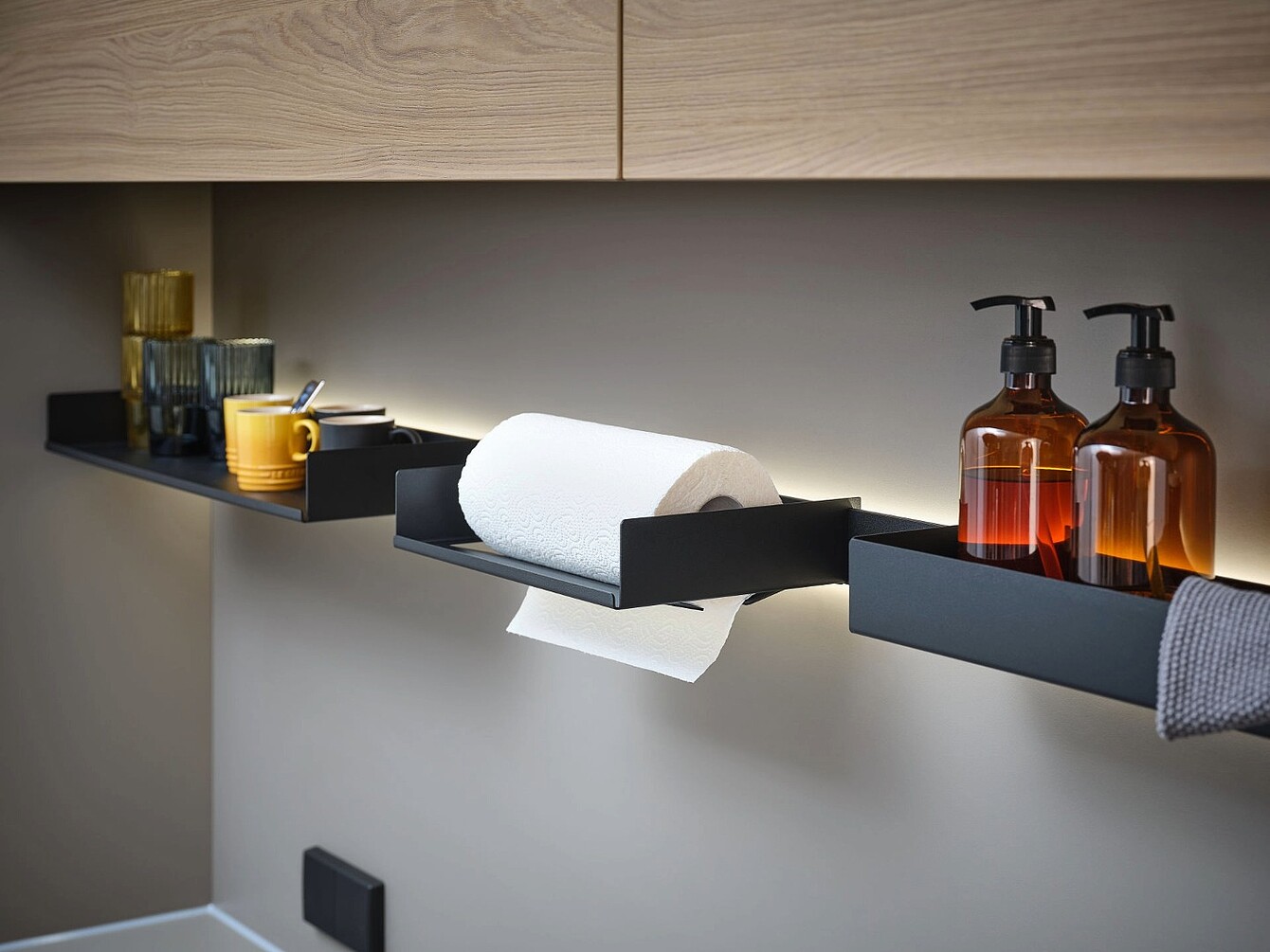 Schwarze Wandregale mit gelben Tassen, Küchenpapierrolle, braunen Seifenspendern und Gläsern vor heller Wand mit indirekter Beleuchtung