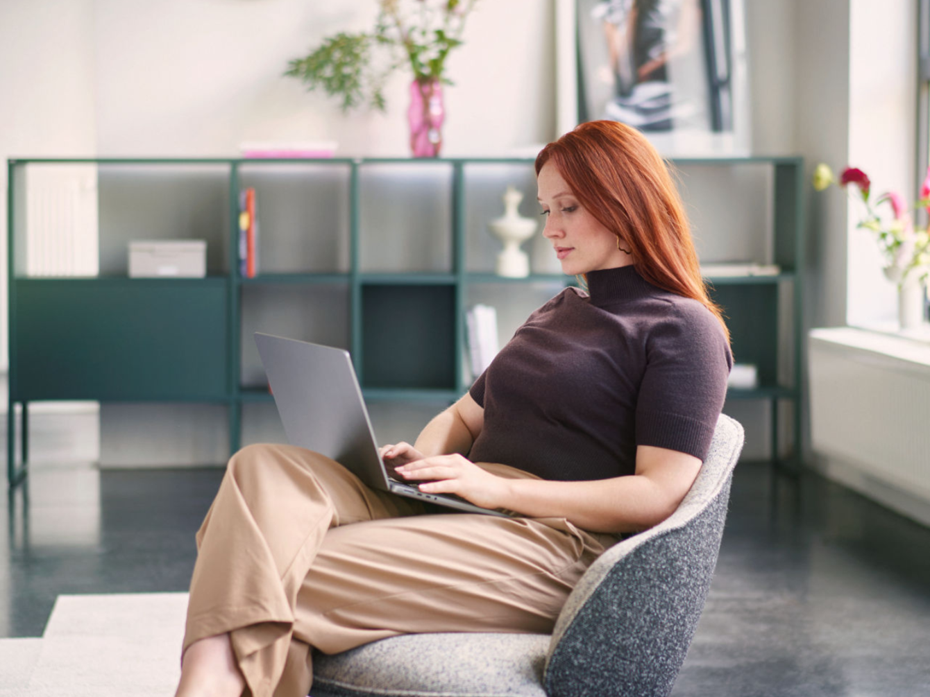 Red-haired woman sitting relaxed in a gray armchair working on a laptop in a modern room with shelves and plants in the background 