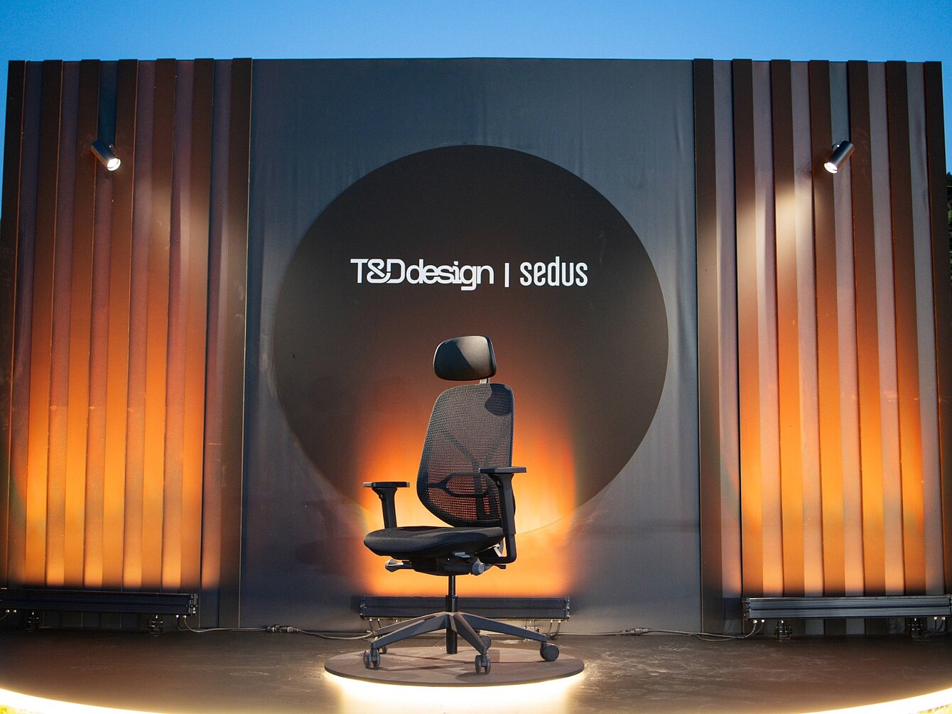 Bürodrehstuhl se:kit mit Kopfstütze vor einer Wand mit beleuchtetem runden Logo 'T&D design | sedus' und vertikalen Holzpaneelen
