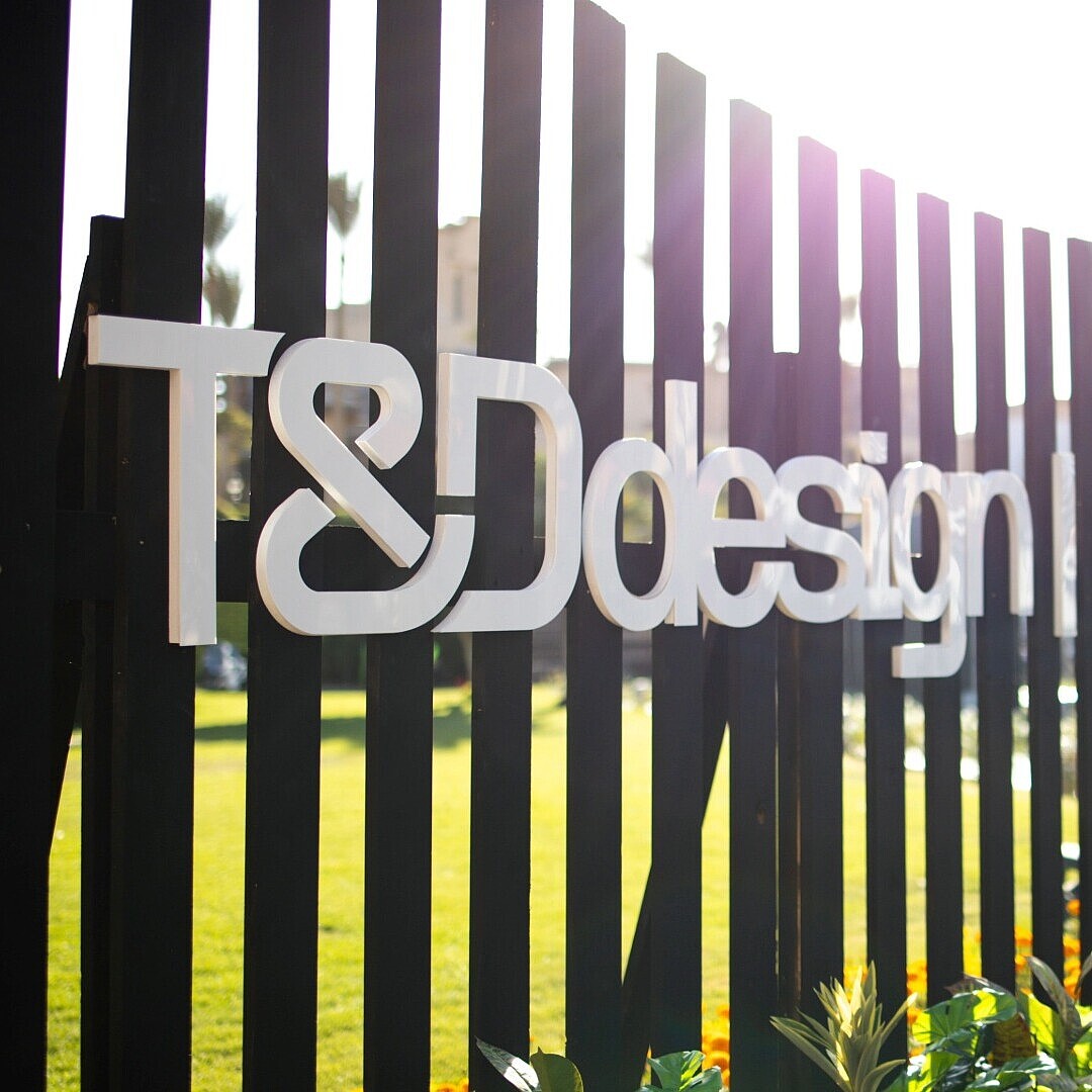 Metallzaun mit vertikalen schwarzen Streben und den weißen Schriftzügen 'T&D design' und 'sedus' im Freien mit Pflanzen im Vordergrund