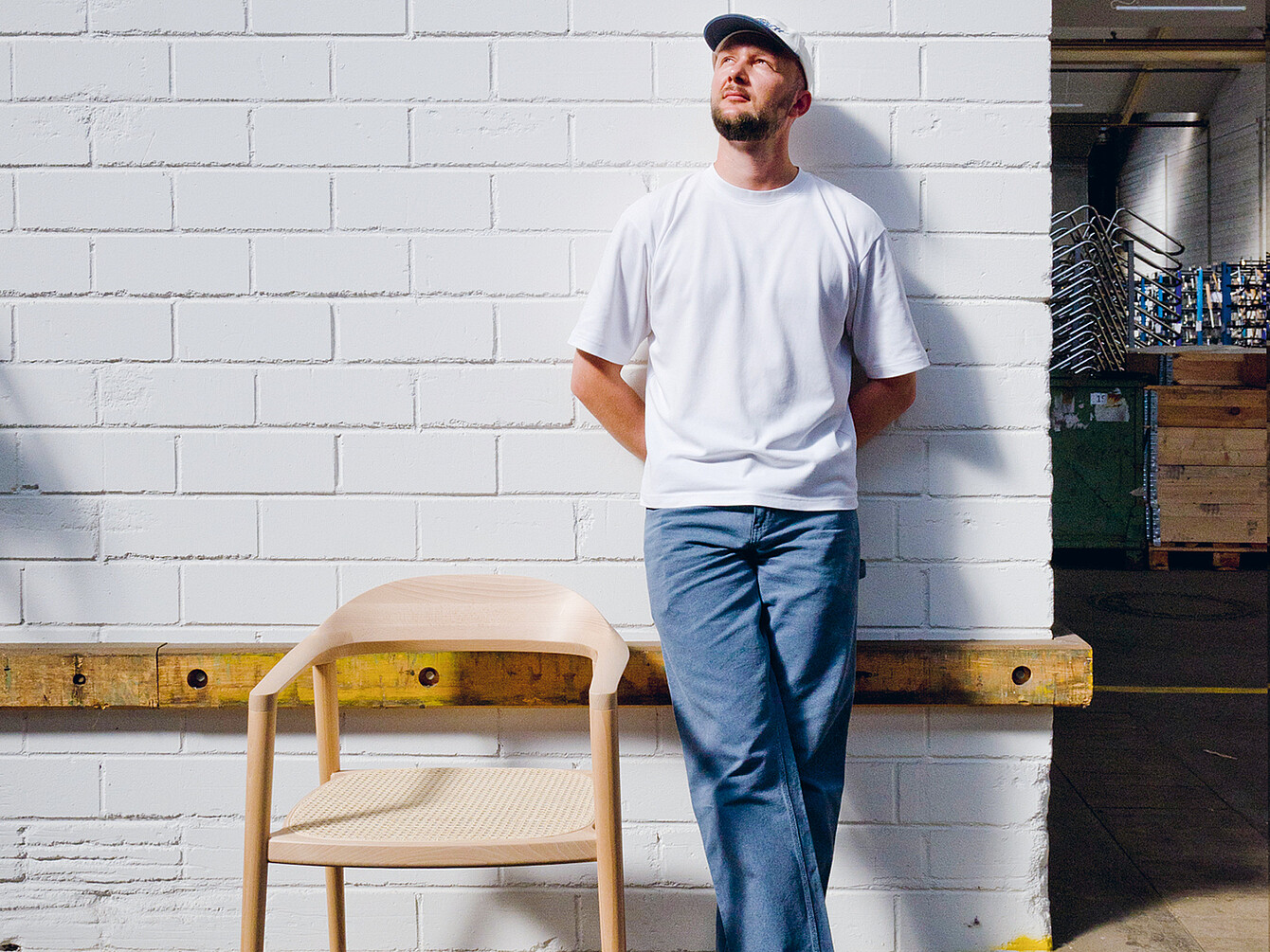Man in casual white shirt and blue pants standing against a white brick wall next to a light se:café wooden chair designed by Konstantin Thomas, highlighting its durable and comfortable craftsmanship.