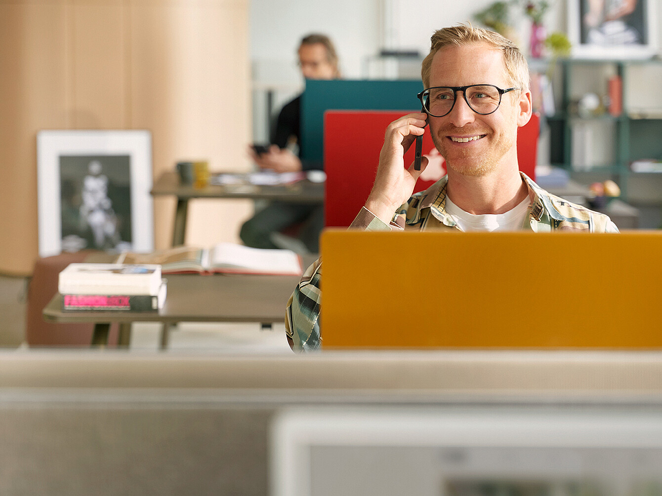 Man with glasses sitting behind a yellow laptop in an office space talking on a phone with a person working at a desk in the background