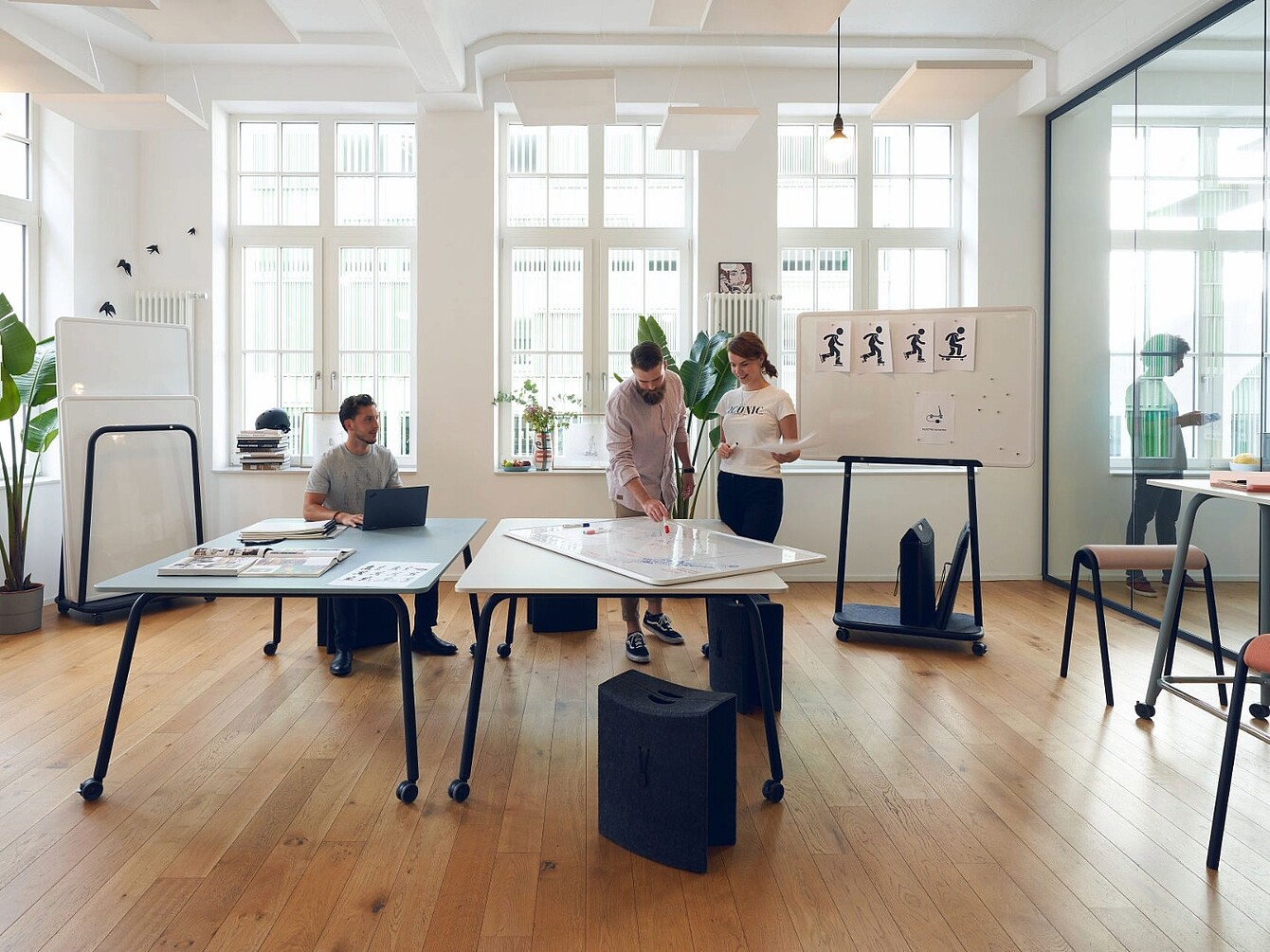 Drei Personen in einem hellen Büro mit großen Fenstern arbeiten gemeinsam an einem Tisch mit Papieren und einem Laptop, im Hintergrund Whiteboards mit Skizzen und Pflanzen