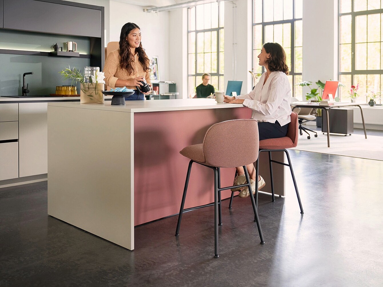 Zwei Frauen unterhalten sich an einer se:café bar Theke mit Hochschränken im Hintergrund in einem hellen, modernen Café oder Büro mit großen Fenstern.