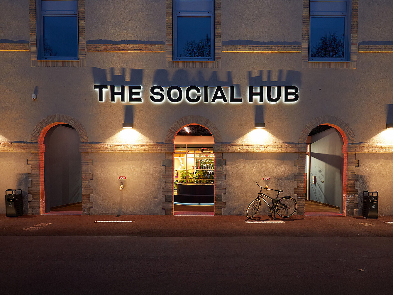 Building facade with three arched entrances lit at dusk with a bicycle parked near one entrance and a sign reading 'THE SOCIAL HUB' above the central arch