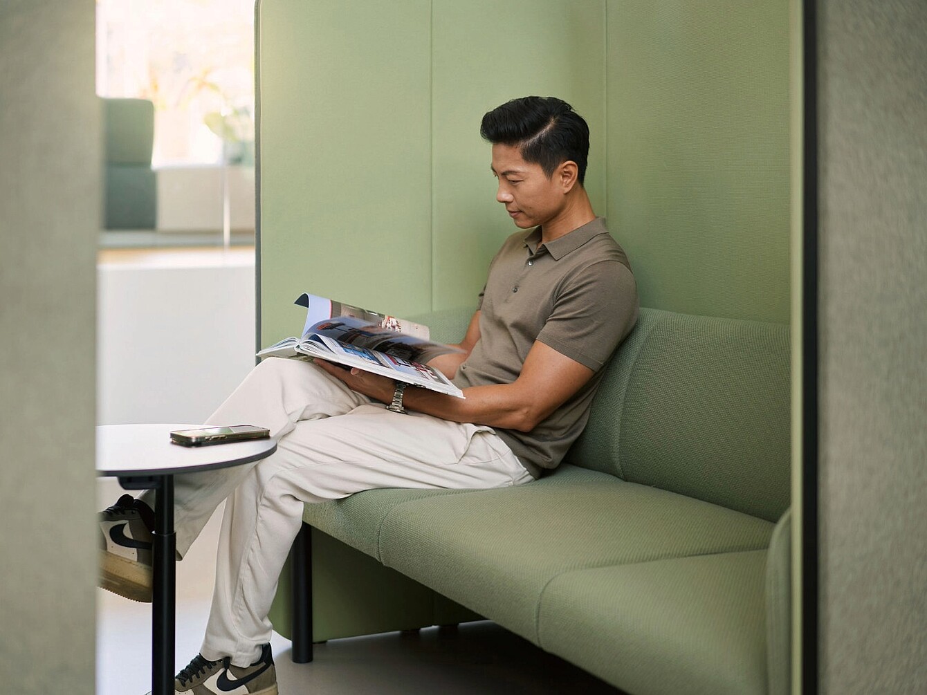 Person sitzt auf grün gepolsterter Bank in einem abgetrennten Raum und liest Magazin