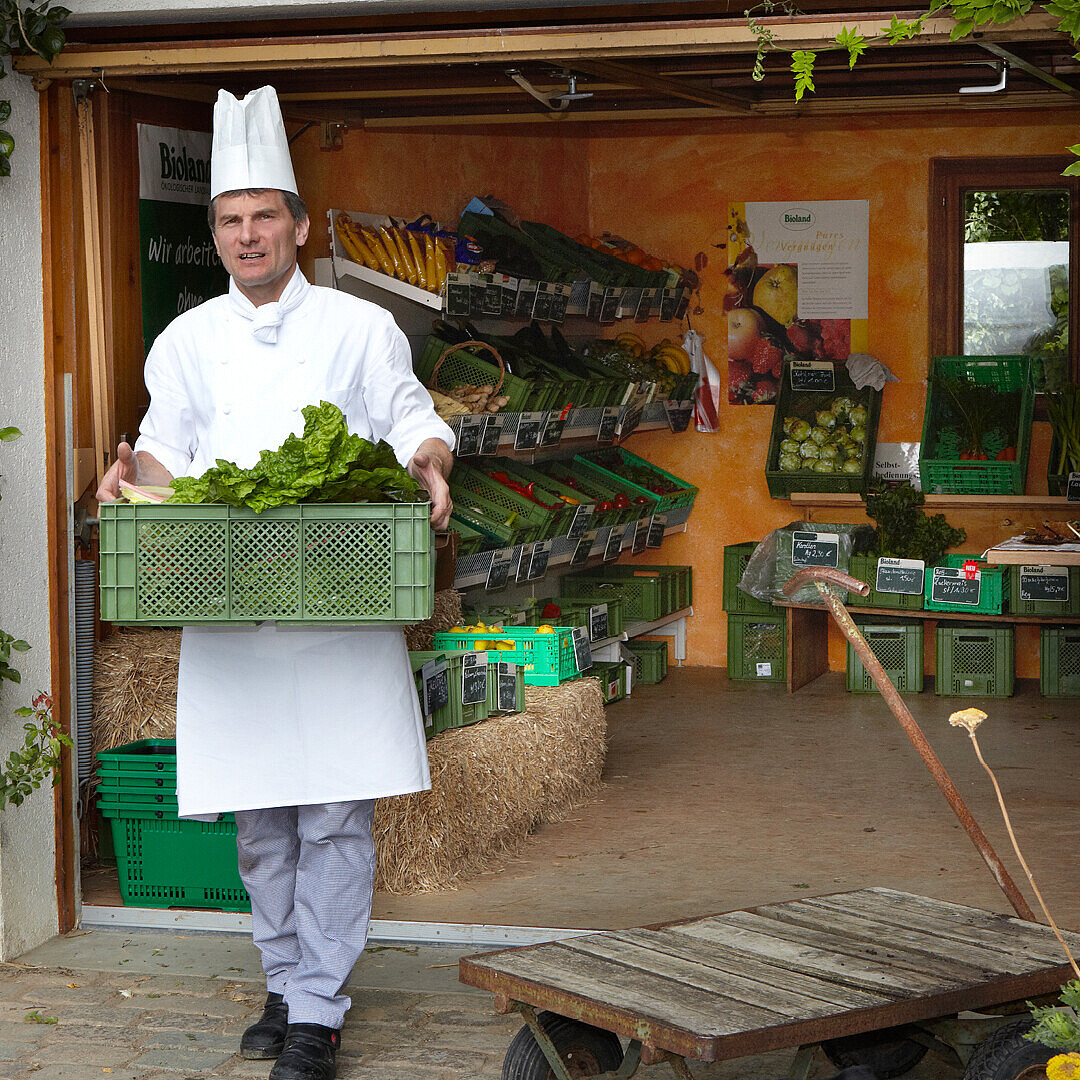 Chef in white uniform holding a crate of fresh lettuce in a rustic farm shop promoting healthy eating and sustainable catering in modern work culture.
