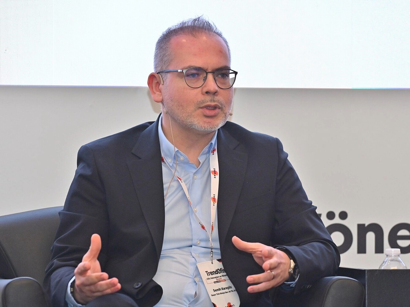 Mann in dunklem Sakko und hellblauem Hemd sitzt auf einem Stuhl und gestikuliert mit beiden Händen vor einem weißen Hintergrund mit teilweise sichtbarem Schriftzug