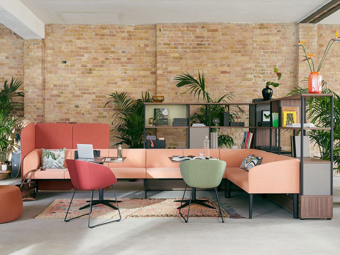 Open lounge area with a peach-colored sectional sofa, two modern chairs in red and green, low tables, plants, and a shelving unit against a brick wall