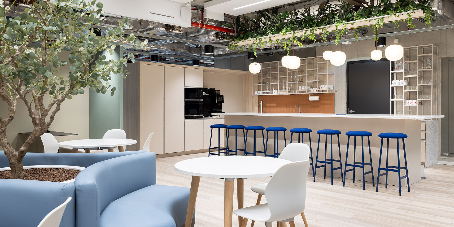 Moderner Bistrobereich bei Veolia mit se:spot Designstühlen in Weiß und blauen Barhockern an se:assist Tischen, umgeben von Pflanzen und heller Beleuchtung. 