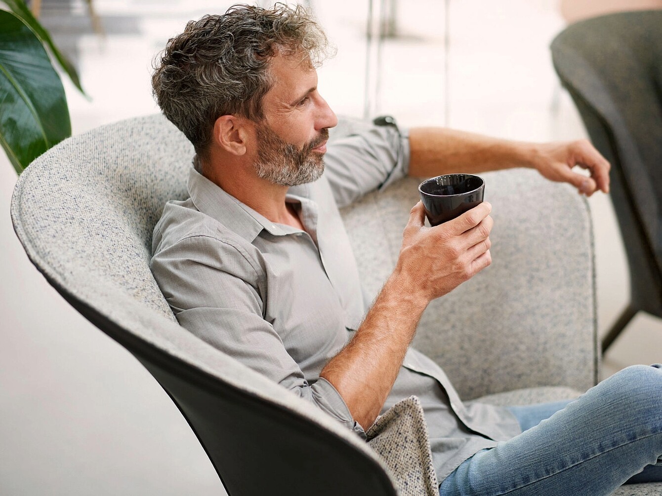 Man with beard sitting in a curved armchair holding a black cup and looking to the side