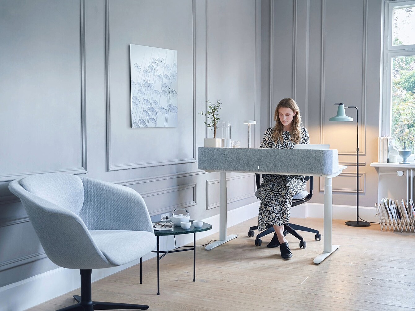 Person sitting at a modern desk in a bright home office with a light armchair, small round table, and wall art