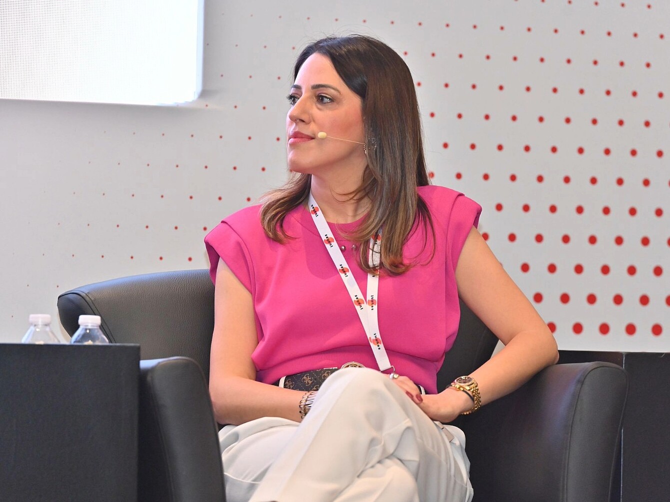 Frau mit schulterlangen braunen Haaren in pinkem Oberteil und hellen Hosen sitzt mit verschränkten Beinen in einem schwarzen Sessel vor einer weißen Wand mit roten Punkten