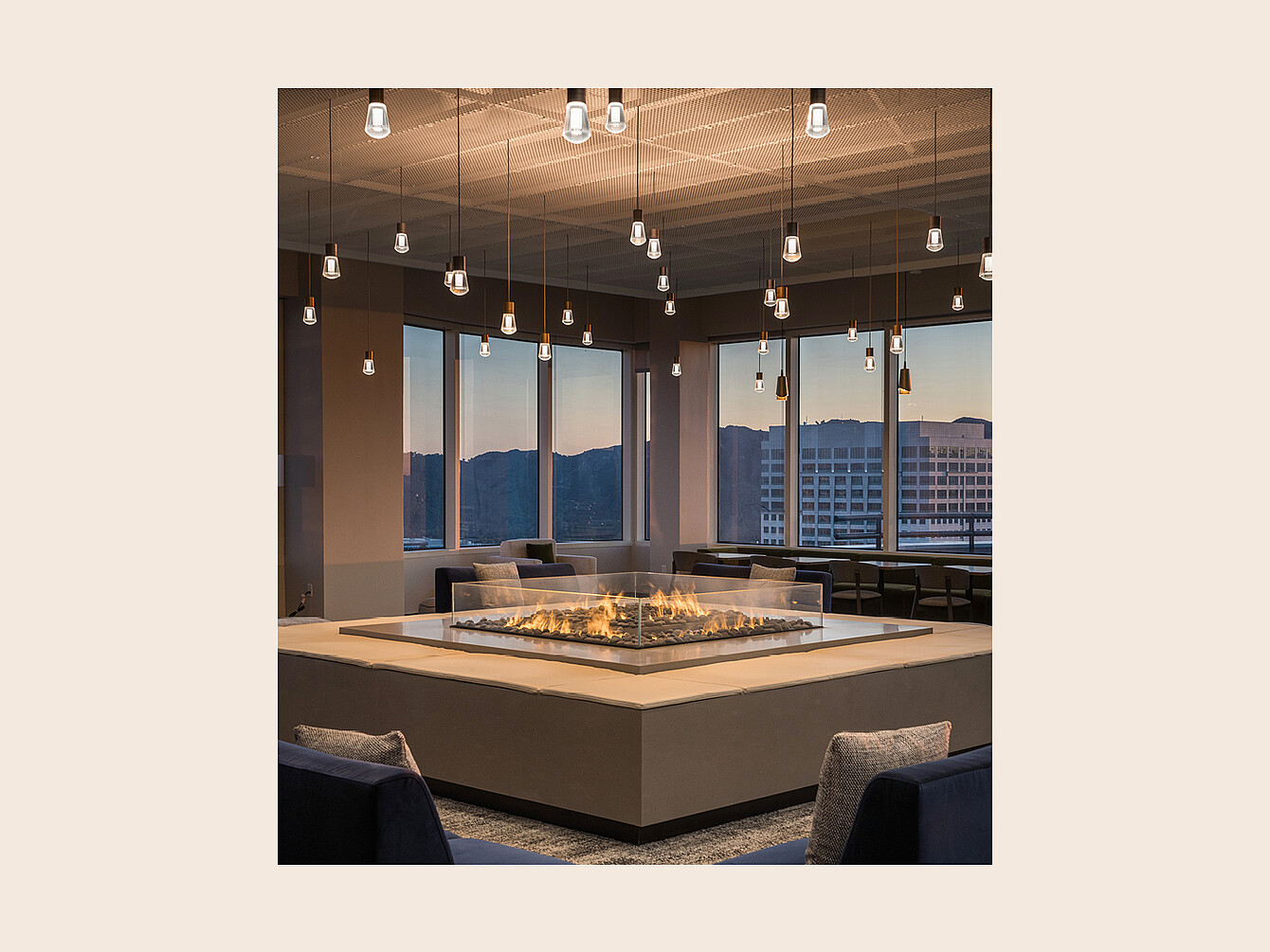 Square recessed fire pit with glass enclosure surrounded by seating in a room with large windows and hanging pendant lights