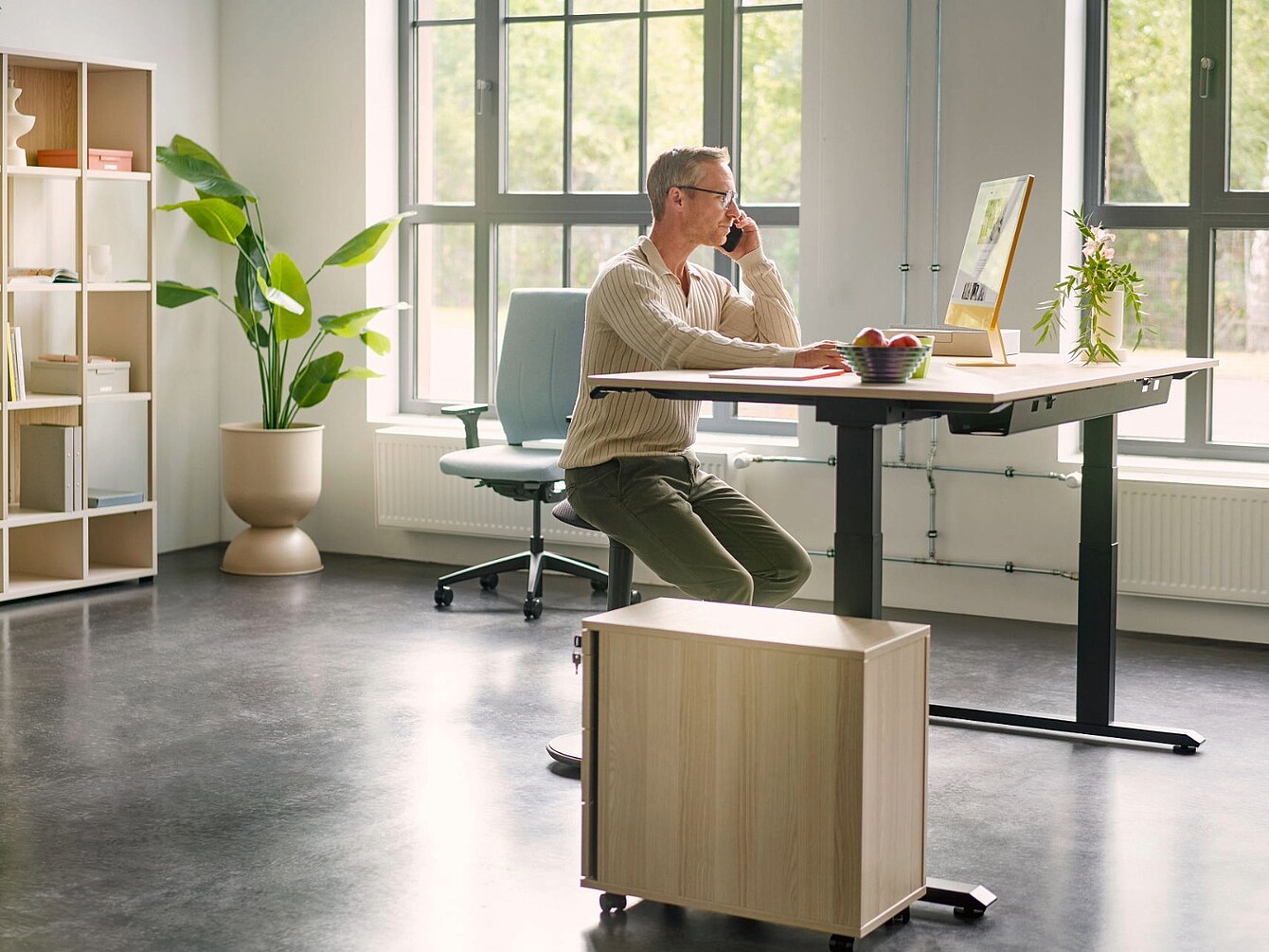 Person sitting on se:fit chair in front of a height-adjustable desk in a bright office with large windows and plants
