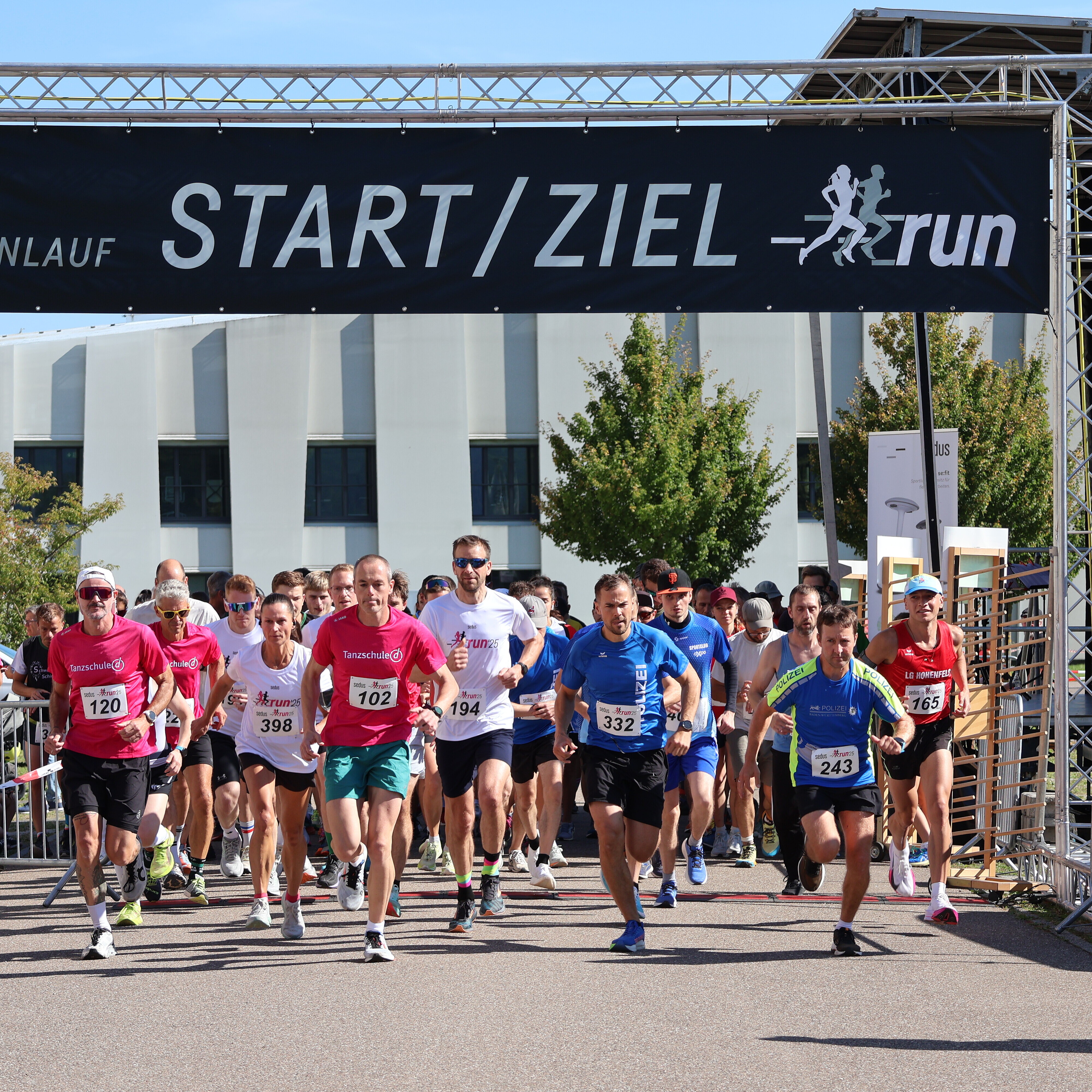 Runners starting the Sedus Firmenlauf run under a black banner reading 'START / ZIEL run' on a sunny day, with participants wearing numbered bibs and athletic clothing.