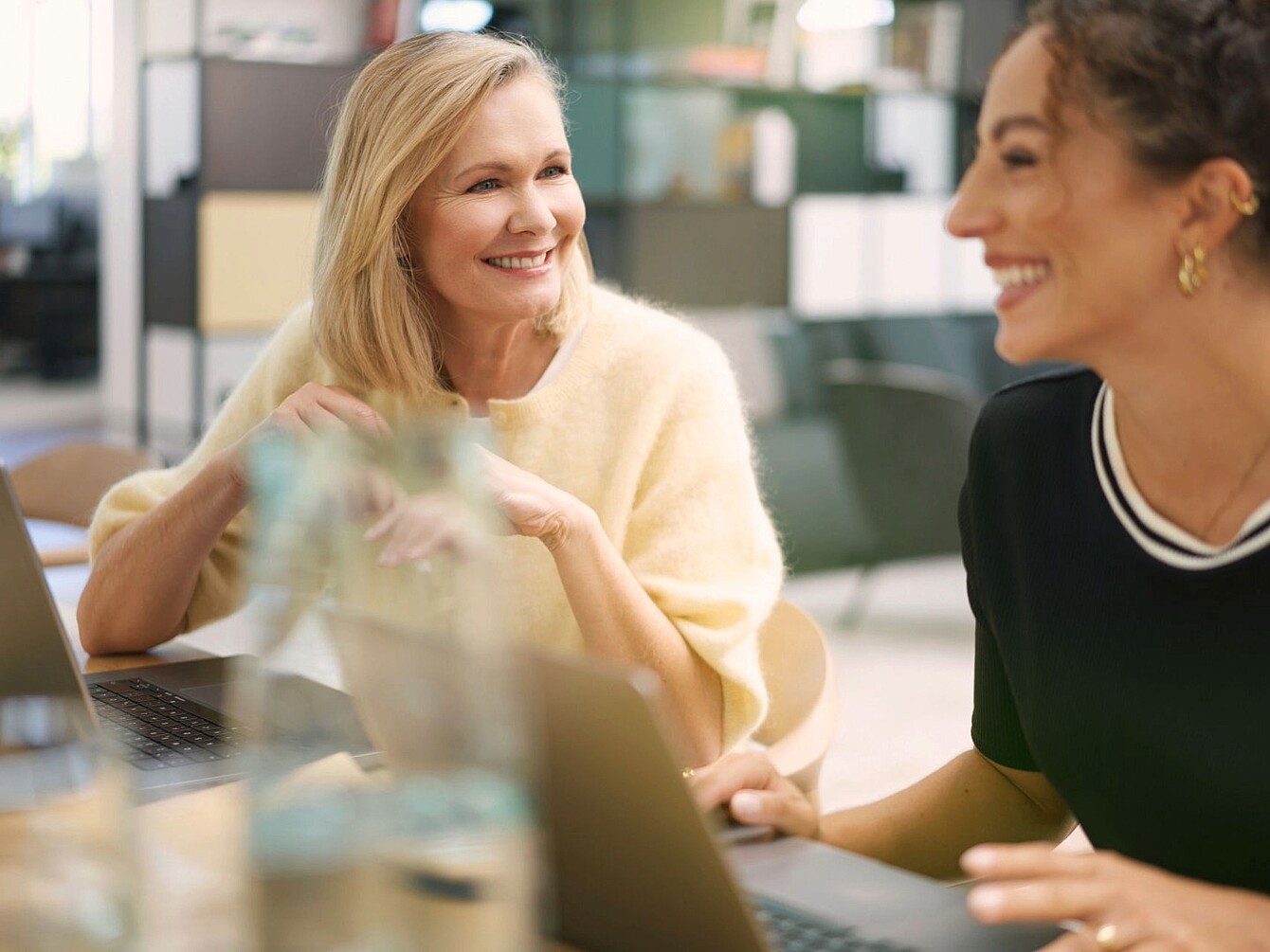 Two women sitting at a table with laptops in a modern office environment, one wearing a light sweater and the other wearing a dark top with white trim and gold hoop earrings