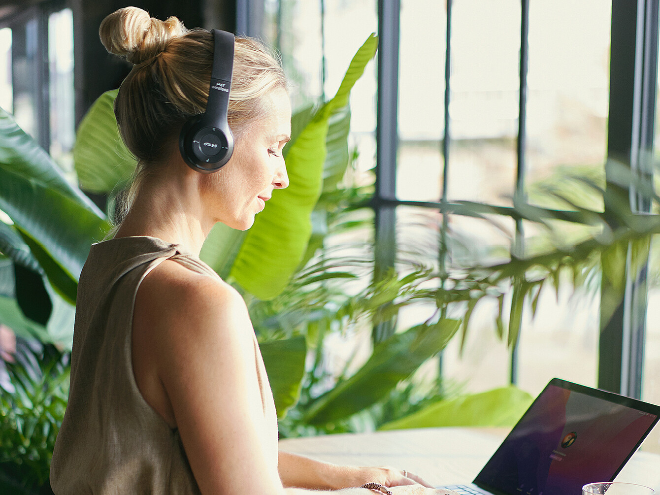 Person wearing headphones working on a laptop in a bright work café surrounded by large green plants and natural light