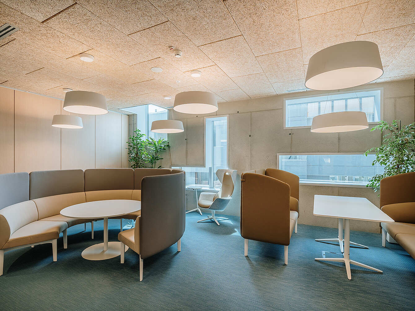 Office lounge area with curved and high-backed seating around round and rectangular tables under large ceiling lights and near windows with plants
