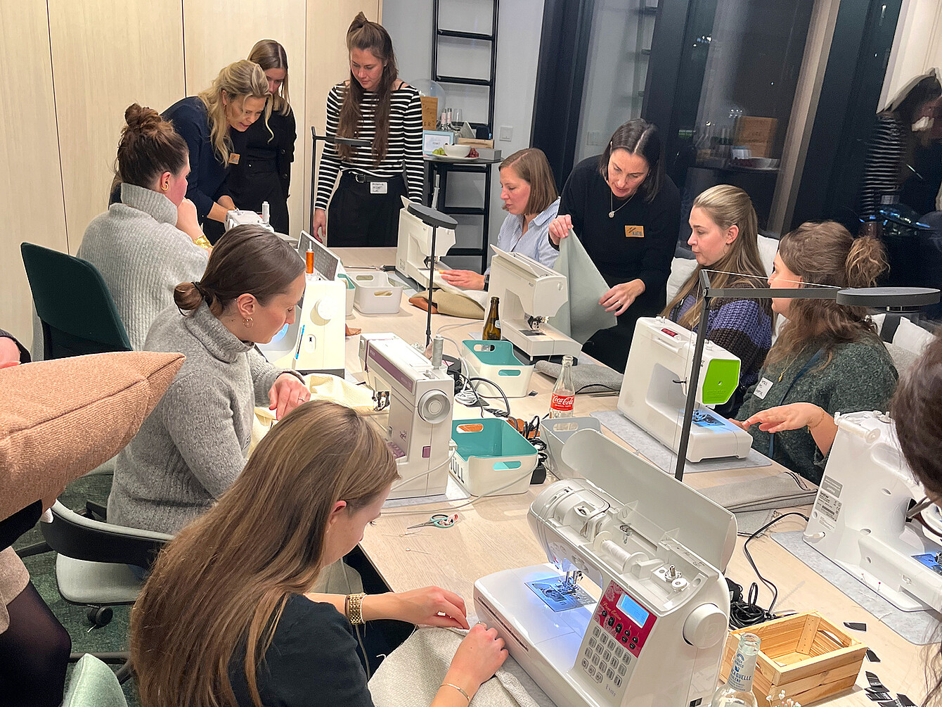 Mehrere Personen sitzen an einem langen Tisch mit mehreren weißen Nähmaschinen, einige stehen und beobachten, im Raum mit großen Fenstern und hellen Wänden.