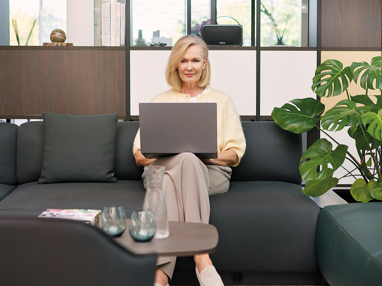 A woman with shoulder-length blonde hair sits on a modern, dark gray se:living sofa by Sedus, holding an open laptop on her lap. She is wearing a light-colored blouse and beige pants. In the background are the se:matrix shelving system from Sedus with decorative items, a large houseplant with distinctive leaves, and a window through which trees can be seen. On the low table in front of her are a water carafe, two glasses, and a magazine.