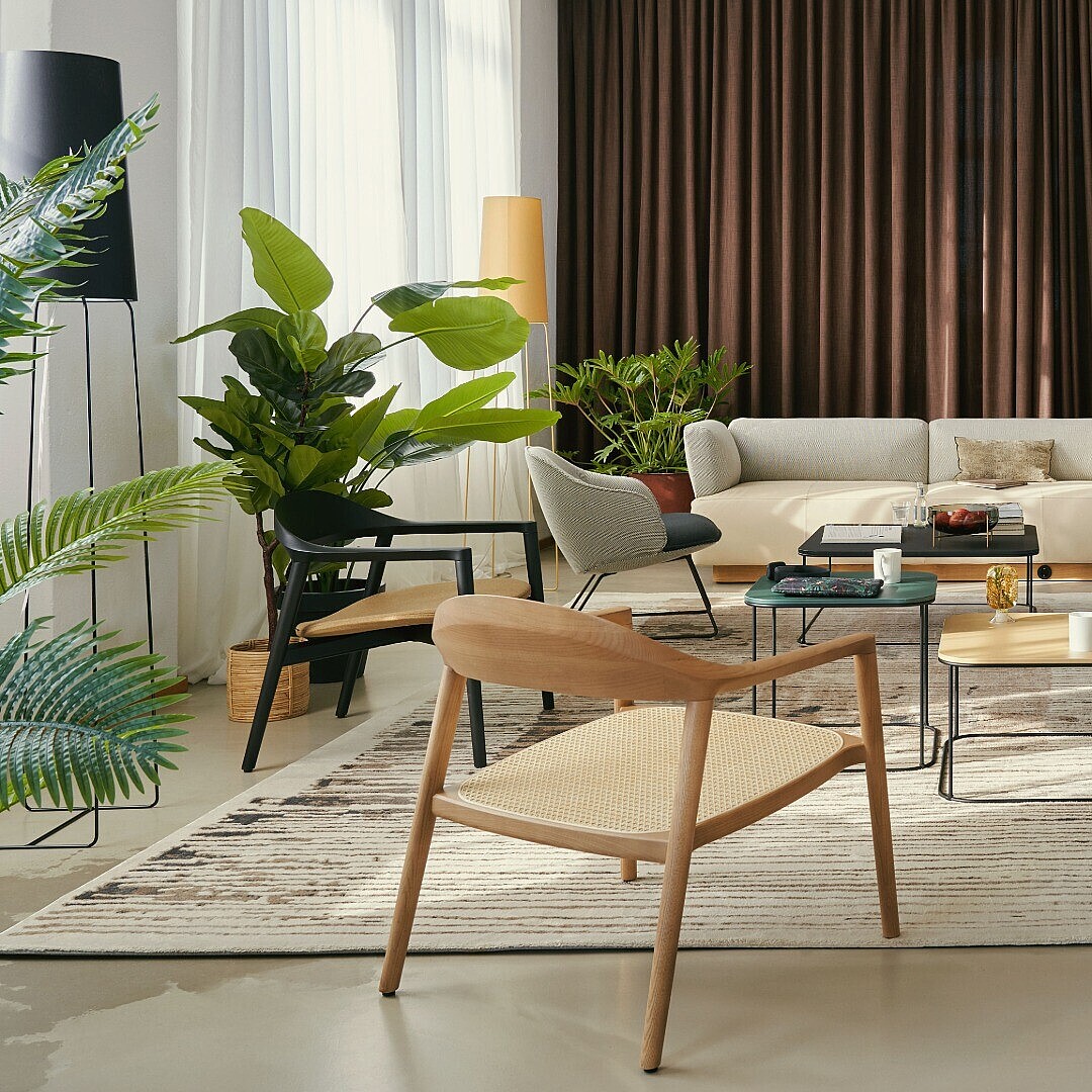 Modern office lounge with multiple plants, wooden and upholstered chairs, low tables, and a sofa against a backdrop of large windows and dark curtains