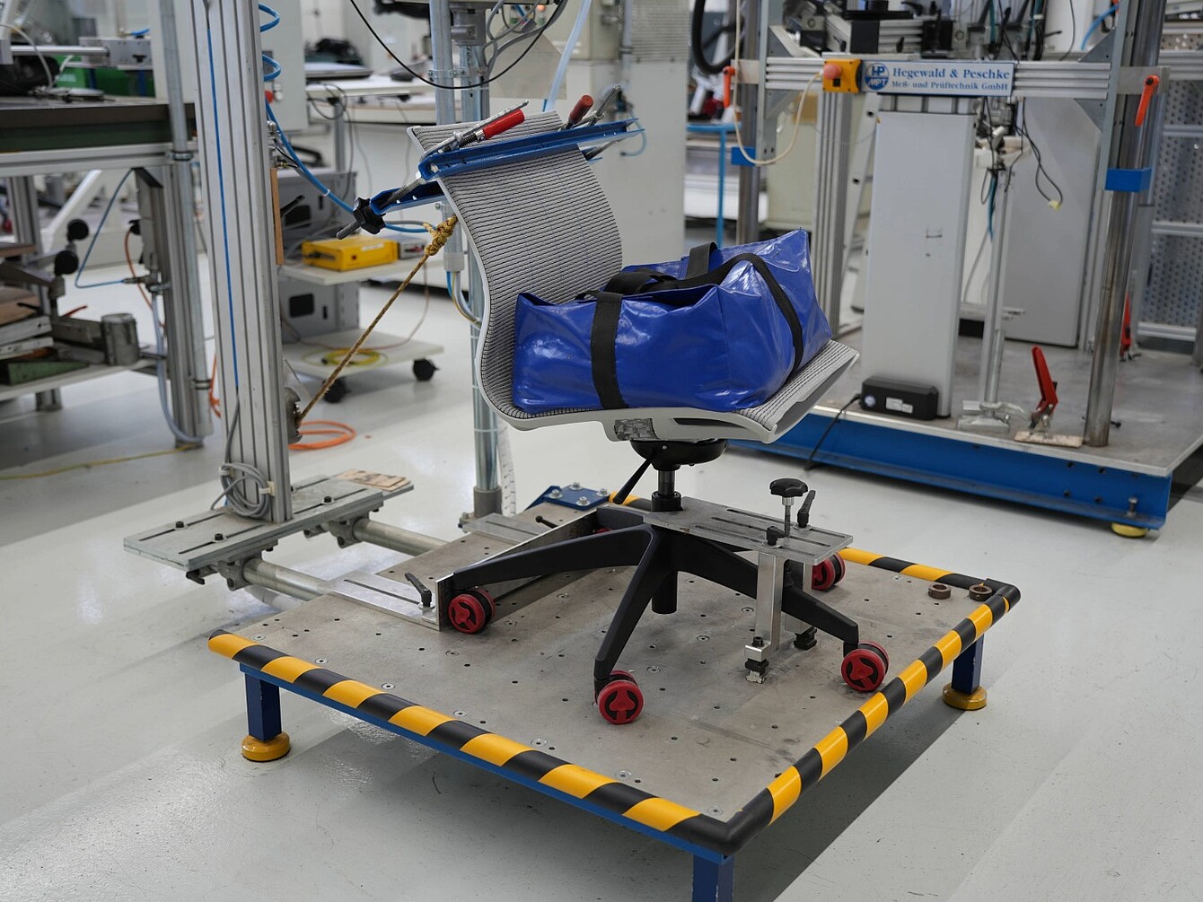Office chair with mesh backrest and blue bag on a test platform with yellow and black striped edges in a laboratory setting