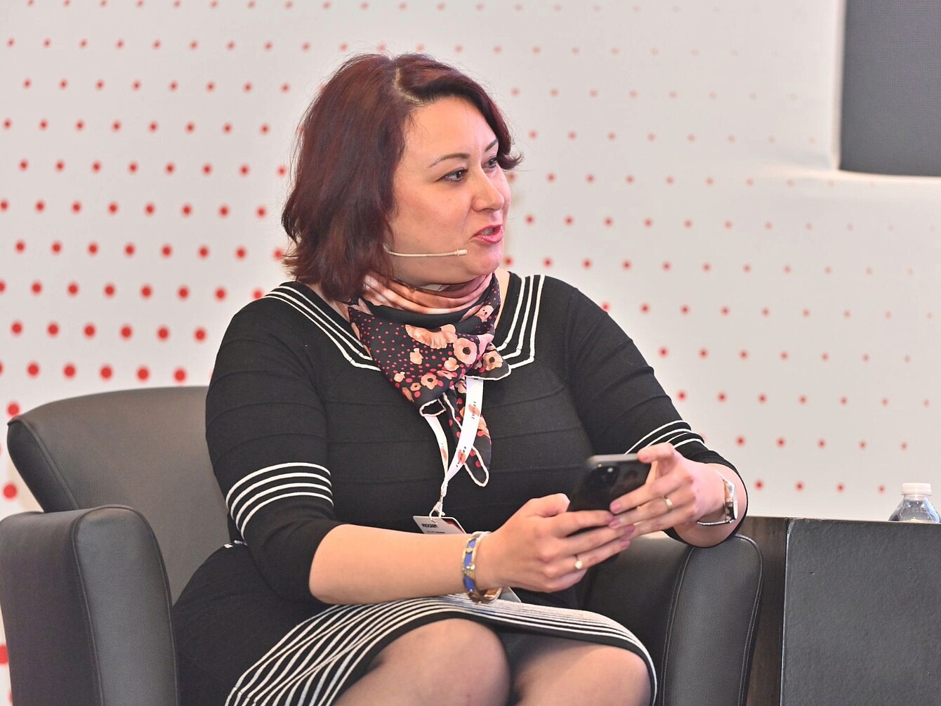 Frau sitzt in einem schwarzen Sessel, trägt ein schwarzes Kleid mit weißen Streifen und ein gemustertes Halstuch, hält ein Smartphone in beiden Händen.