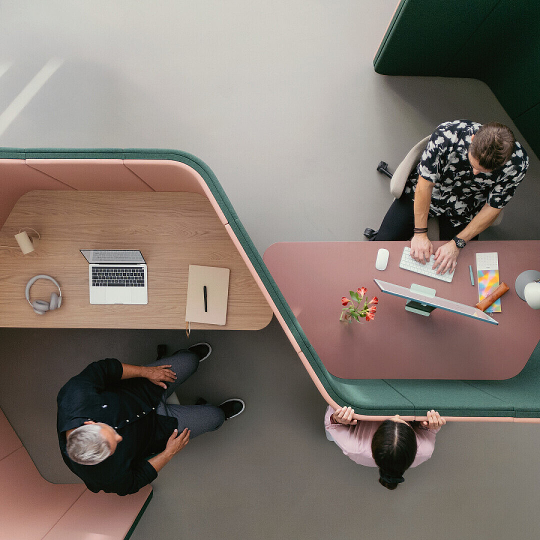 Overhead view of two specialized workspaces with hexagonal green and pink padded walls; one workspace has a person sitting with a laptop and headphones, the other has a person working on a desktop computer with a colleague standing nearby, illustrating a modern office environment supporting focused and collaborative work.