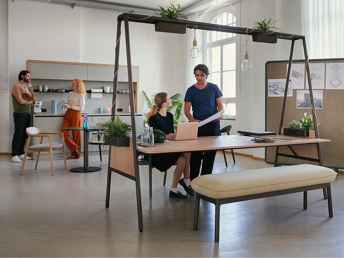 Modern hybrid ductile commons workspace with two people collaborating at a wooden table with plants and hanging lights, and two others chatting near a kitchenette in the background