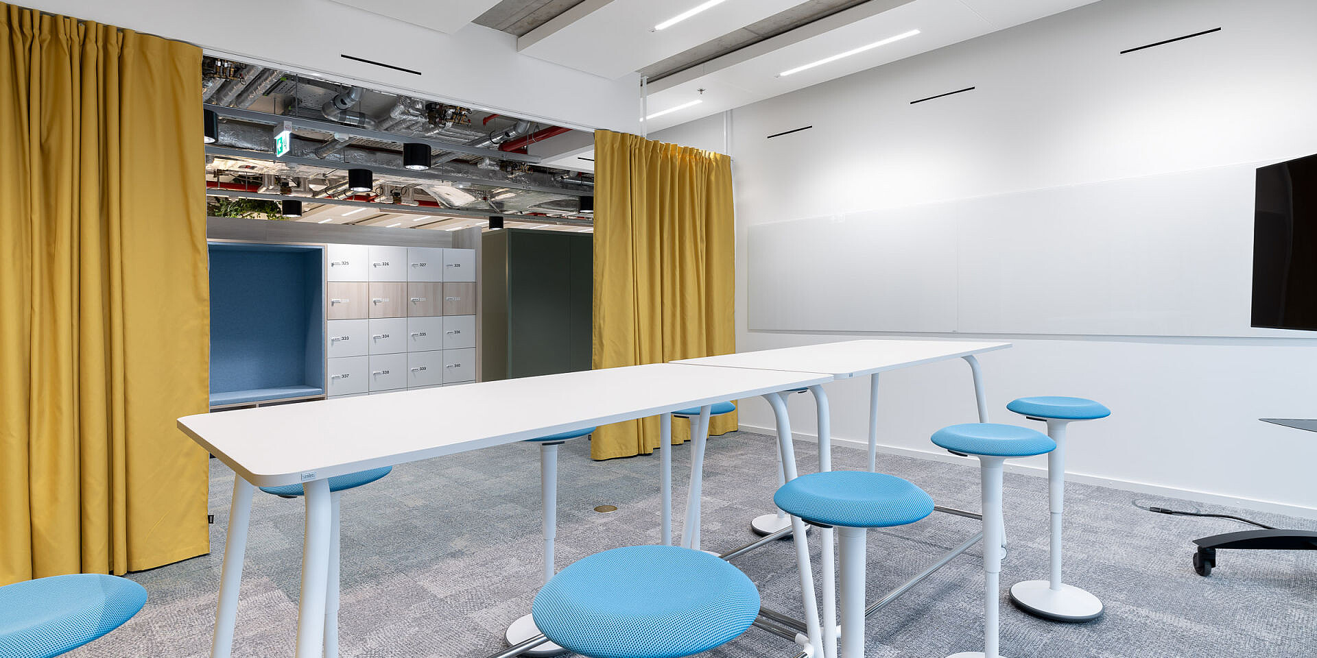 Moderner Besprechungsraum mit weißem se:lab high desk Workshop-Tisch, umgeben von blauen se:fit Stehsitzen und se:dot Hockern, gelbe Vorhänge und weiße Wände. 