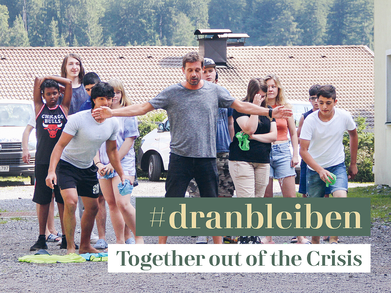 Group of young people outdoors, some stretching and others standing, with a building and parked cars in the background, overlaid with text '#dranbleiben Together out of the Crisis'.
