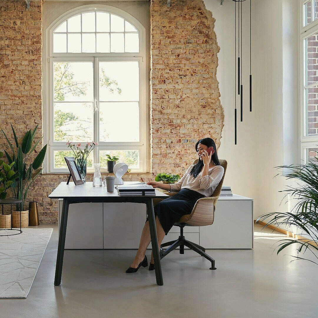 Person sitting at a desk in a modern office with exposed brick walls, large window, plants, and lounge seating area