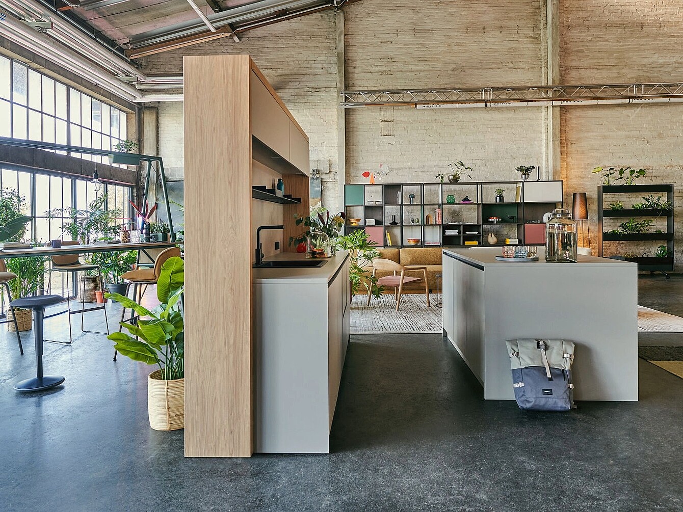 Offene se:café bar aus hellem Holz, moderner Kücheninsel und Sitzbereich mit Pflanzen in einem loftartigen Raum mit hohen Fenstern und sichtbaren Deckenbalken
