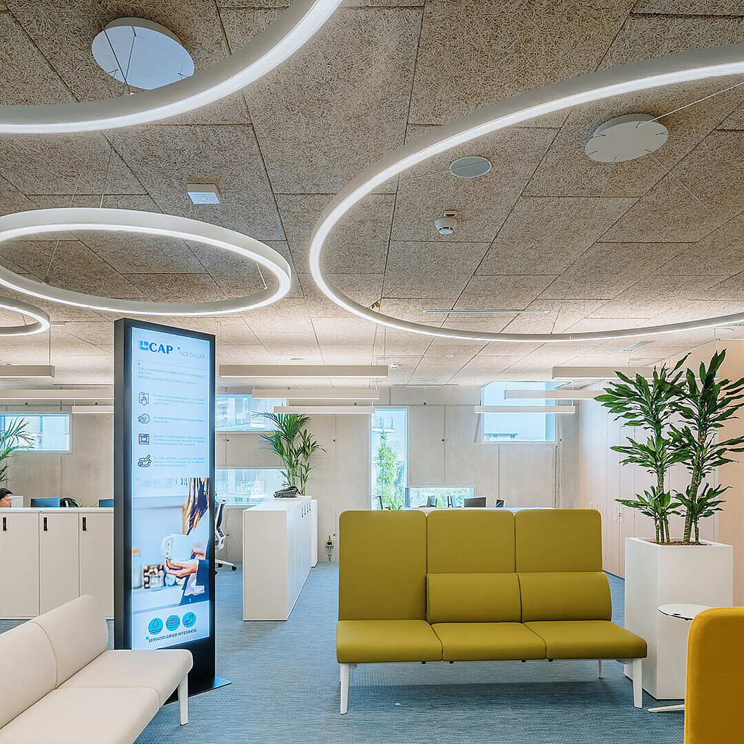 Open-plan office space with white and yellow sofas, large potted plants, circular ceiling lights, and a digital information display