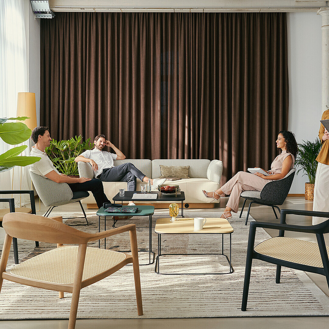 Moderne Business Lounge mit bequemen Sesseln, Sofas und Pflanzen, in der vier Personen entspannt arbeiten und sich unterhalten