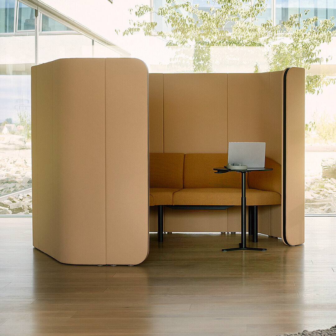 Secluded seating booth with a sofa, small table and laptop in a bright, modern office environment.