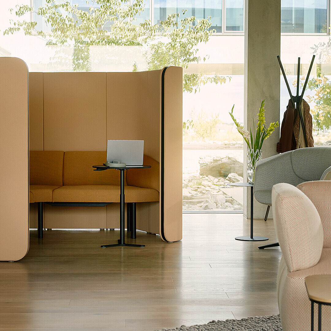 Abgeschirmte Sitznische mit Sofa, kleinem Tisch und Laptop in einer hellen, modernen Büroumgebung.