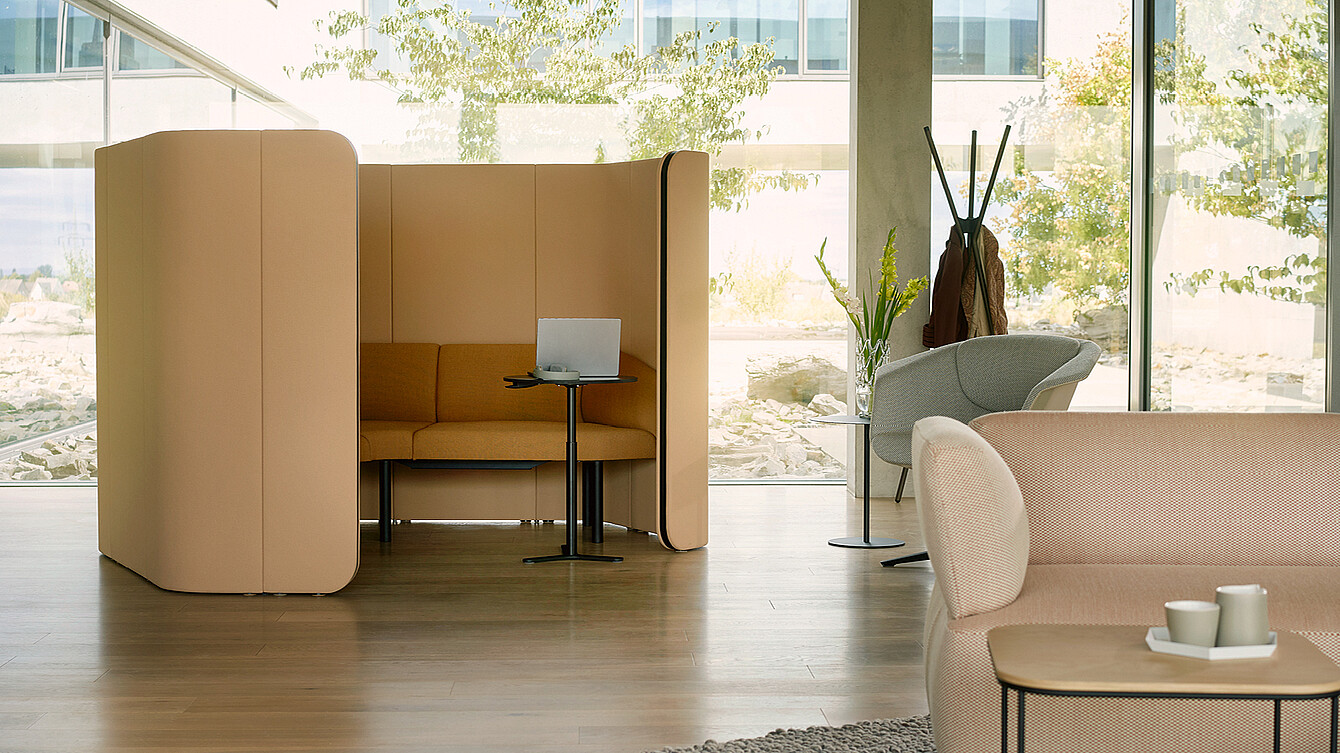 Abgeschirmte Sitznische mit hoher Rücken- und Seitenwand, Sofa, kleinem Tisch und Laptop in einer hellen Büroumgebung.