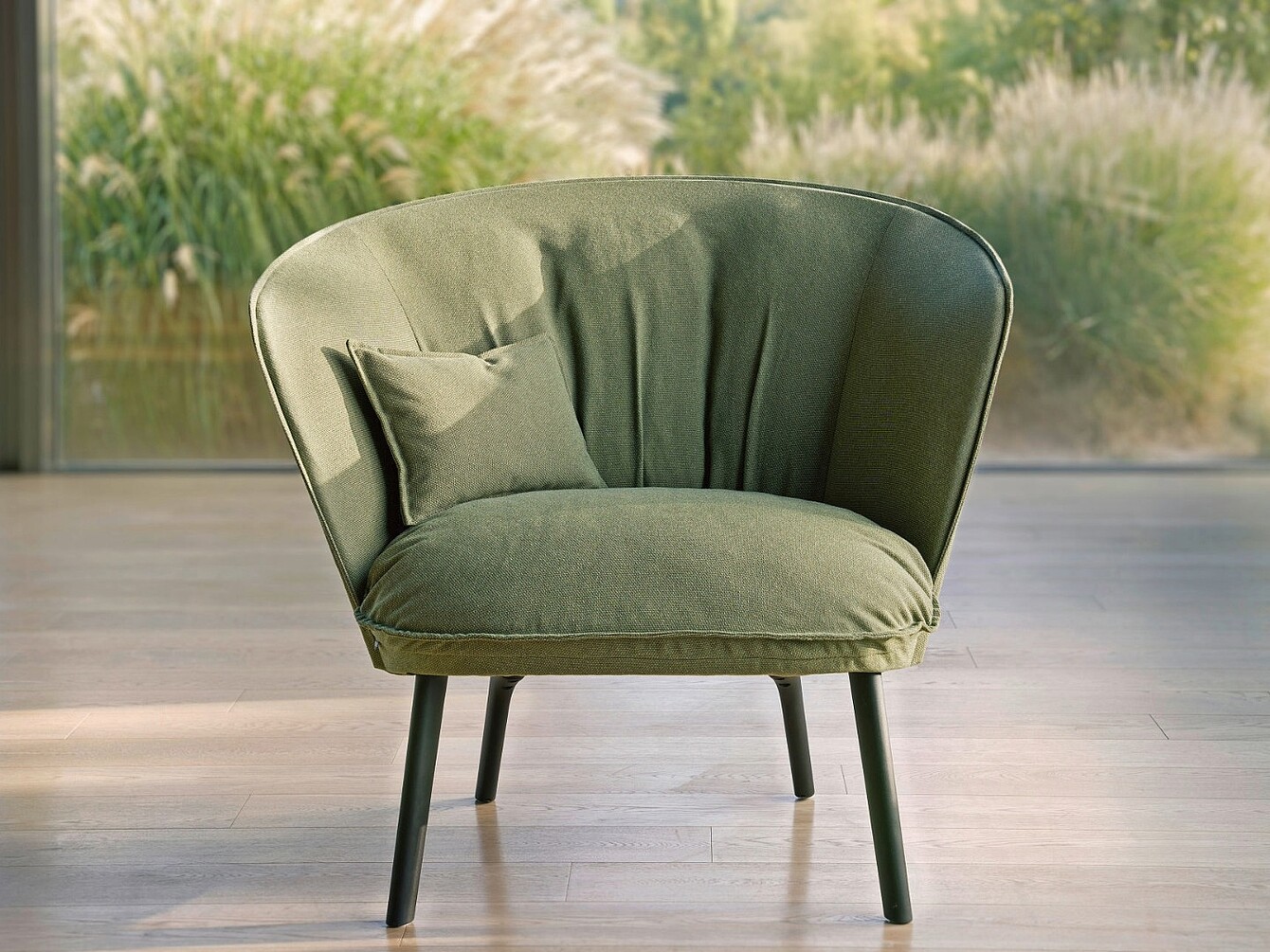 Moderner, gepolsterter Sessel mit abgerundeter Rückenlehne und kleinem Kissen vor großem Fenster mit Blick auf Pflanzen