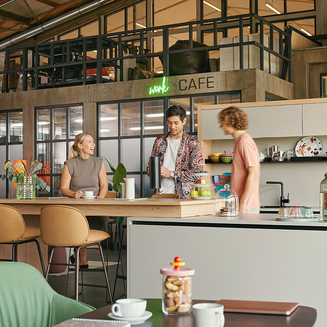 Three people standing and sitting around a modern kitchen island in a flexible work café space with bar stools, plants, and open shelving