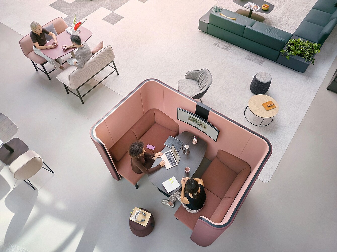 Zwei Personen sitzen in einer halbhohen, gepolsterten, rosa-farbenen Rückzugszone an einem Tisch mit Laptop und Notizbuch, umgeben von weiteren Sitzgelegenheiten in einem offenen, hellen Raum.