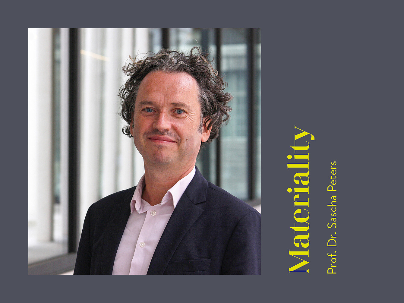 Man with curly hair wearing a dark blazer and light shirt standing indoors near large windows with vertical frames