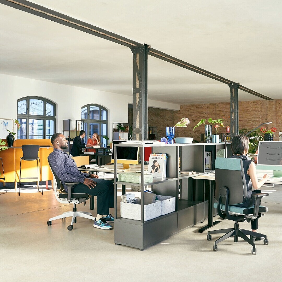 Großzügiger, offener Büroarbeitsplatz mit mehreren Personen an Schreibtischen, modernen Bürostühlen, Pflanzen und großen Fenstern in einem Raum mit Sichtbacksteinwand und Stahlträgern.