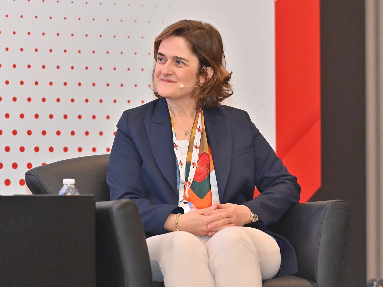 Frau sitzt mit verschränkten Händen auf einem schwarzen Sessel, trägt dunkelblauen Blazer, weiße Hose und bunt gemusterten Schal, im Hintergrund rote und weiße Wandgestaltung