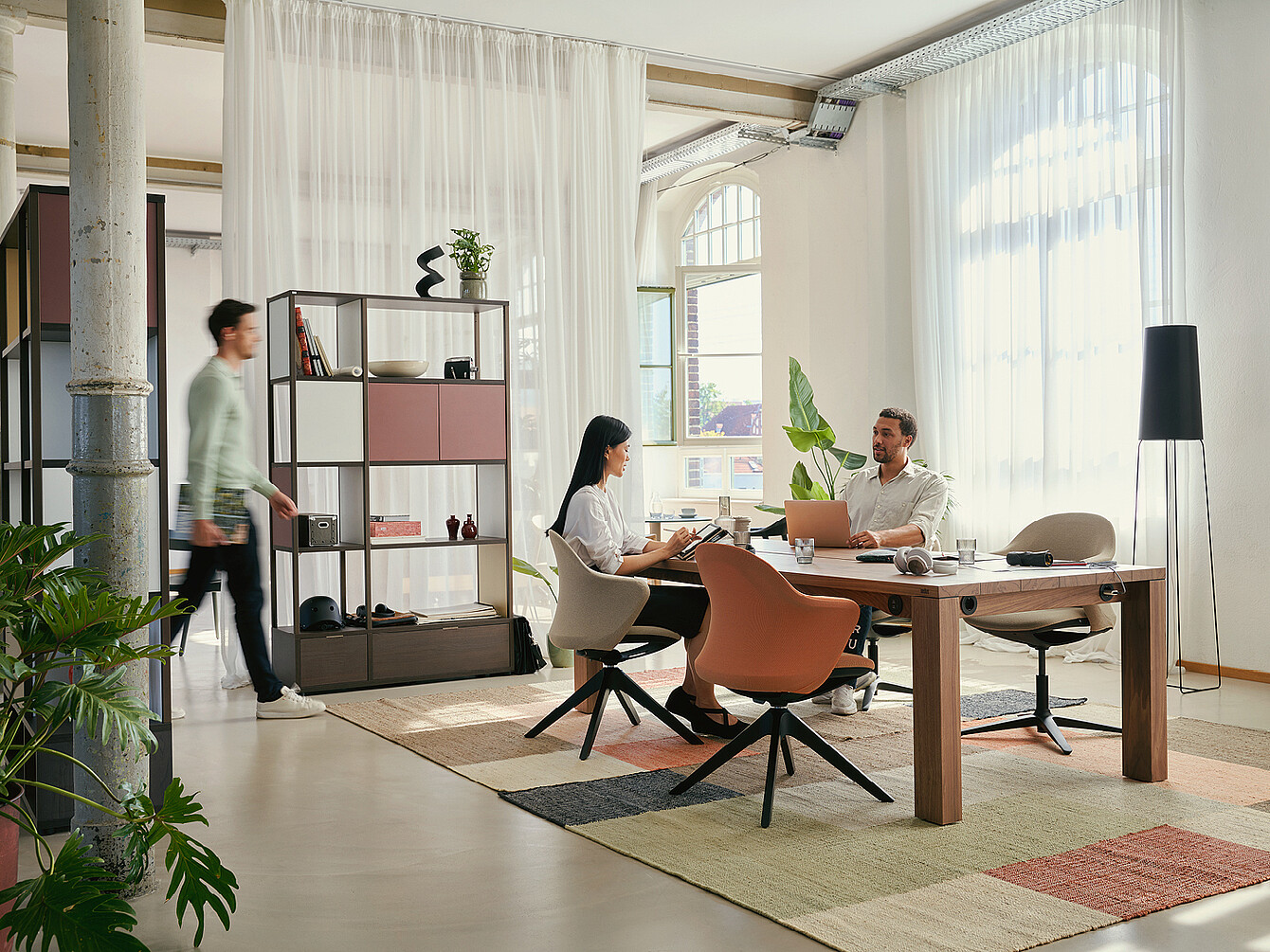 Modern hybrid ductile commons workspace with two people seated at a wooden table using laptop and tablet, surrounded by plants, shelves, and large windows with sheer curtains, emphasizing flexibility and community.