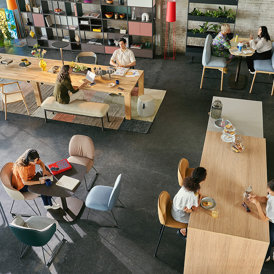 Employees working and socializing in a work café with diverse seating areas including long tables, small round tables, and lounge chairs, illustrating varied atmospheres for different tasks and moods.