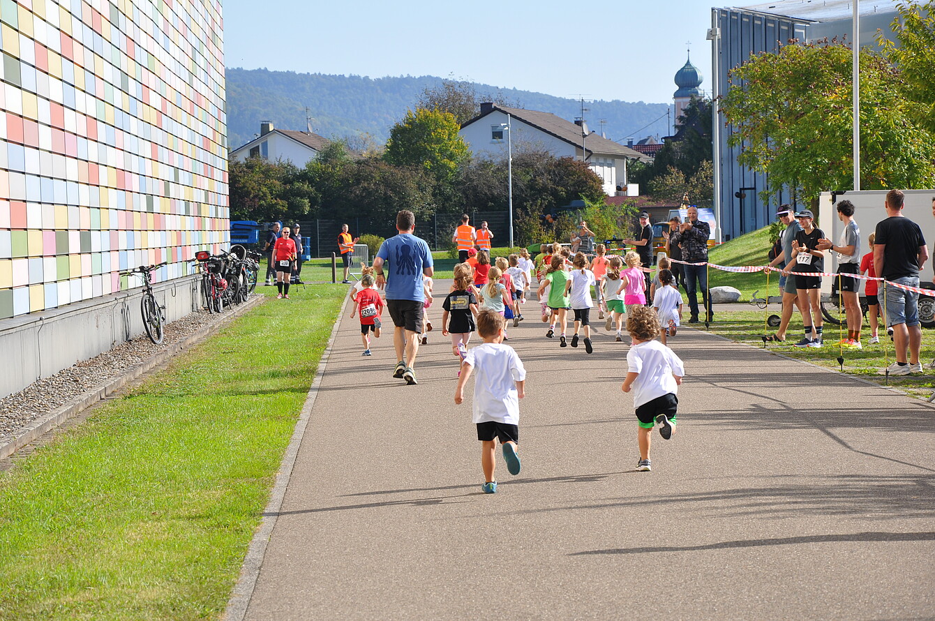 Children running on a paved path during the Sedus RUN Kids event, with adults and spectators along the sides and a colorful building on the left.