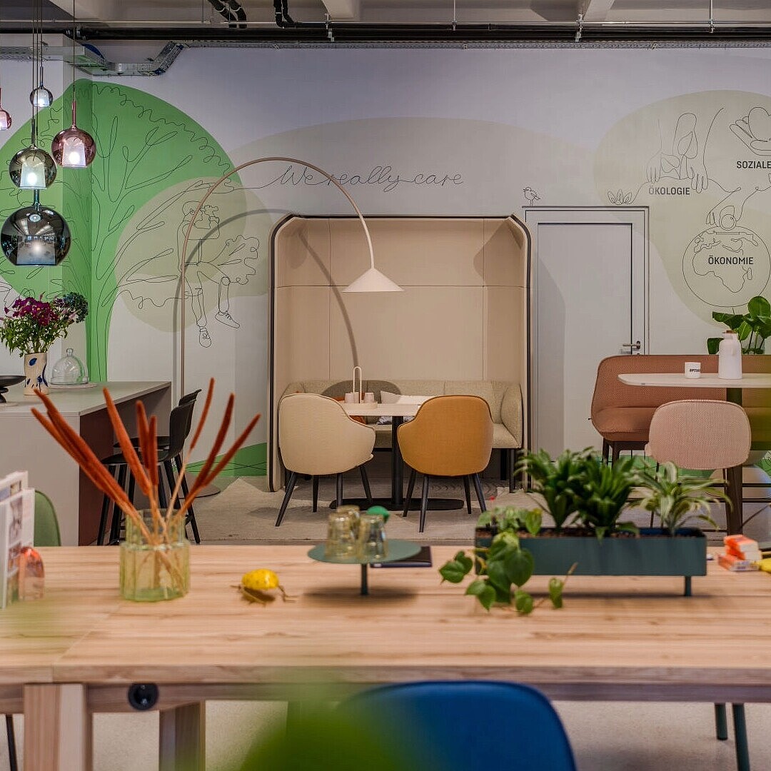 Moderner, heller Arbeitsbereich mit großem Holztisch, verschiedenen Stühlen, Pflanzen und einer Sitznische im Hintergrund