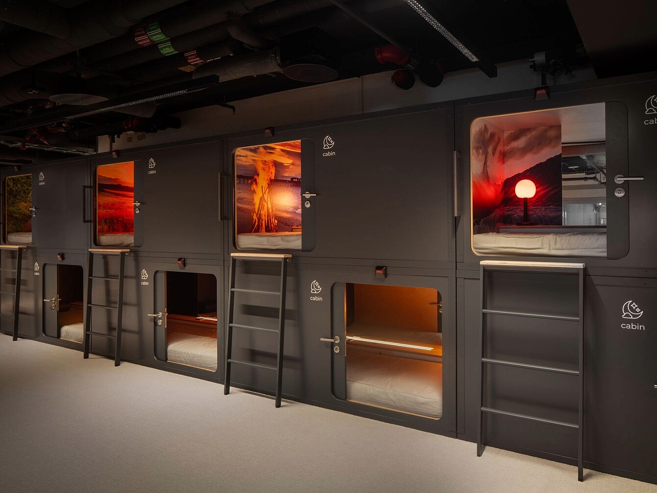 Mehrere zweistöckige, schwarze Schlafkabinen mit Leitern und großen Fenstern, die Bilder von Sonnenuntergängen zeigen, im Novu Campus am Züricher Flughafen.